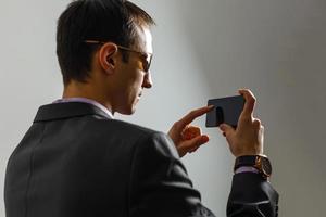 Young cheerful brunete lawyer is standing on the pure light blue background and smiling, reading good news on internet at his mobile phone, wearing suit photo