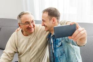 Happy moment. Cheerful young man taking a selfie with his upbeat elderly father waving at the camera and smiling pleasantly photo