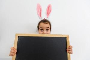 niña jugando al bebé en la búsqueda de huevos de pascua juego de niños. tablero de tiza en blanco con forma de corazón para su texto foto