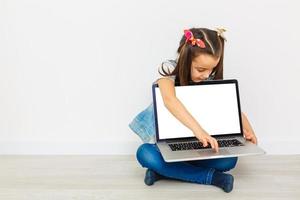 una niñita linda está sentada en el suelo con su laptop, usando anteojos, aislada en blanco foto