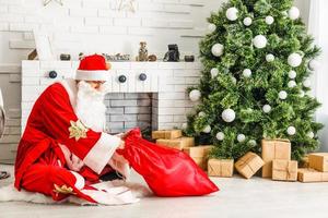 Santa Claus sitting in front of fireplace near Christmas tree with a bag full of presents and a wish list photo