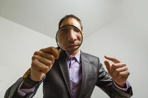 young male detective holding magnifying glass by right eye while standing in front of camera in isolation photo