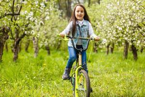 Child riding a bike on street with blooming cherry trees in the suburbs. Kid biking outdoors in urban park. Little girl on bicycle. Healthy preschool children summer activity. Kids play outside photo