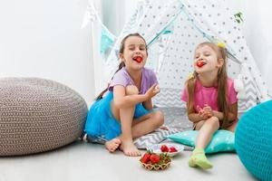 two little girls eat strawberries at home near wigwam tent photo