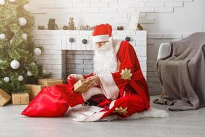 Santa Claus sitting in front of fireplace near Christmas tree with a bag full of presents and a wish list photo