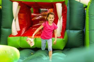 linda y graciosa niña preescolar en un colorido, saltando y rebotando en un castillo inflable divirtiéndose en una fiesta de cumpleaños para niños en un parque infantil en verano foto