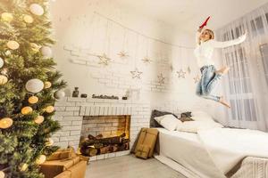 encantadora chica vestida con suéter blanco y pantalones salta sobre la cama con manta gris y almohadas blancas en una acogedora habitación decorada con un árbol de año nuevo foto