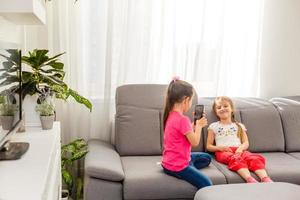 people, children, technology, friends and friendship concept - happy little girls sitting on sofa and taking selfie with smartphone at home. photo