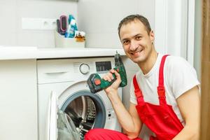 Young attractive smiling worker in uniform fixing washing machine, background photo