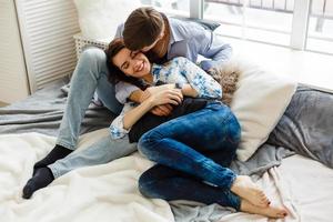Stylish hipster couple of man and woman sitting on bed in their home in cozy casual clothes. They hug and love each other and smile happily, resting together. Space for text photo