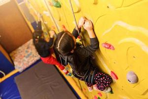 Little baby girl with funny hear style climbing vertical wall and man belaying her from below photo