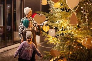 tres niños divirtiéndose cerca del árbol de navidad iluminado al aire libre por la noche. foto