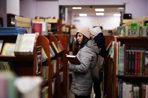 Mother with son in jacket and backpack reaching a book from bookshelf at the library. photo