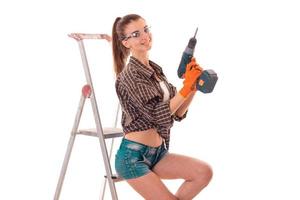 sexy brunette lady makes refurbish an apartment with drill in hands and ladder isolated on white background in studio photo