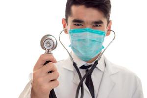 close up of young male doctor in uniform and mask with stathoscope posing isolated on white background photo