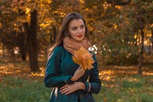 dulce niña se encuentra en el parque de otoño mira lejos y mantiene las hojas en la mano foto