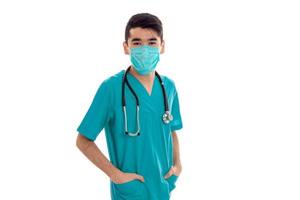 young handsome brunette man doctor in uniform and mask with stethoscope on his shoulders looking at the camera isolated on white background photo