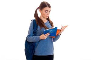Una joven y hermosa estudiante con mochila lee un libro y posa aislada de fondo blanco en el estudio foto