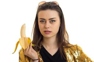 Charming brunette woman in golden jacket eats a banana and looks at the camera photo