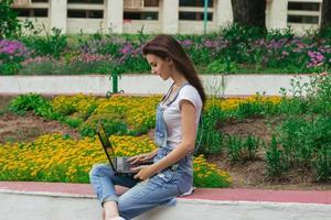 cute young girl sitting near flower beds with flowers and looking at laptop photo