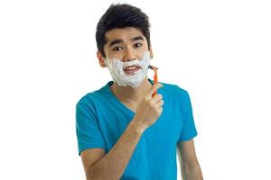 cute young guy in a blue t-shirt and with black hair looks straight and shaves his beard machine with foam on his face photo