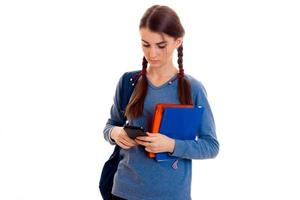 young beautiful student girl with backpack looking at her mobile phone and posing isolated on white background in studio photo