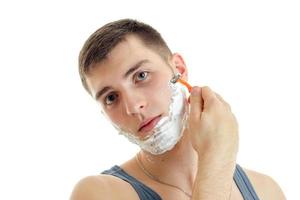 young guy with foam on his face looks into the camera and shaves his beard machine photo