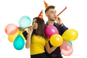 feliz pareja joven soplando cuernos en una fiesta de cumpleaños con globos en sus manos foto