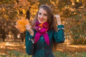 hermosa niña sonriente en una bufanda rosa se encuentra en el parque y mantiene las hojas foto