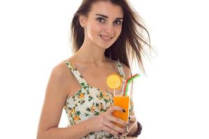 happy young brunette woman in sarafan with floral pattern smiling on camera and drinks orange cocktail isolated on white background photo