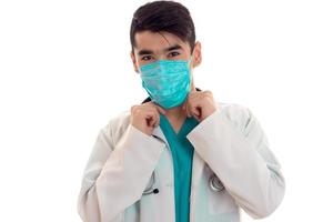 joven médico apuesto posando en uniforme y máscara aislado en fondo blanco en el estudio foto