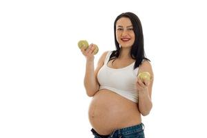 Una alegre morena embarazada con una gran barriga posando con manzanas verdes en las manos aisladas de fondo blanco foto