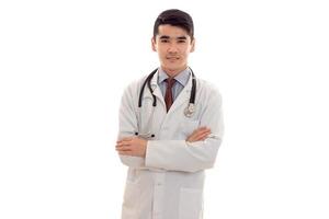 handsome young brunette man doctor in white uniform with stethoscope looking and smiling on camera isolated in studio photo