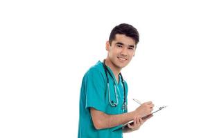 young cheerful brunette man doctor in blue uniform with stethoscope on his shoulders make notes and smiling on camera isolated on white background photo