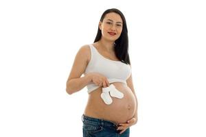 joven embarazada con el pelo oscuro tocando su gran barriga con pequeños calcetines blancos en las manos y mirando la cámara aislada en el fondo blanco foto