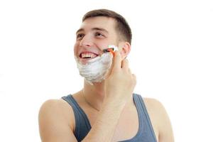 a cheerful young man with foam on his face looks toward the machine and shaves his beard