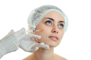 una joven sin maquillaje con la gorra médica levantó los ojos y la cosmetóloga la pincha en el primer plano de la cara foto