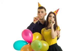 Una pareja joven y divertida celebra su cumpleaños con conos en la cabeza, mantienen bolas de colores y soplan cuernos foto