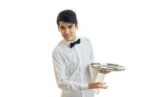 atractivo joven camarero con una camisa blanca sonriendo inclinándose hacia adelante y sosteniendo una bandeja foto