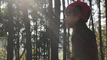 Adorable little girl in warm clothes and red knit hat sits hugging herself on a wooden chair in a pine forest in winter with sunlight. video