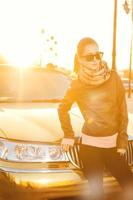 Beautiful adult woman in leather jacket and scarf in golden sunlight near car photo