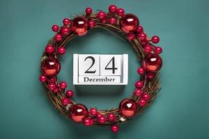 Christmas wreath decorated with red berries, wooden calendar date 24 December isolated on green background Concept of Christmas preparation, atmosphere Wishes card Hand made Christmas wreath Flat lay photo