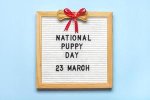 Felt board with text National puppy day in 23 march, dog accessories - bone with red bow on blue background Top view Flat lay Holiday card photo