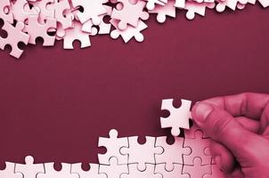 The hand folds a white jigsaw puzzle and a pile of uncombed puzzle pieces lies against the background of surface. Texture photo with space for text Image toned in Viva Magenta, color of the 2023 year