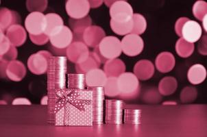 Small red gift box and golden coin stacks on the colored bokeh background Image toned in Viva Magenta, color of the 2023 year photo