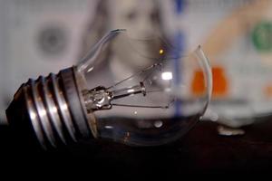 Broken light bulb with pieces of glass on 100 dollar bills  blur background. Concept of electricity prices and the electricity crisis in World photo