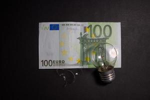 bombilla rota con trozos de vidrio en el billete de 100 euros. concepto de precios de la electricidad y la crisis de la electricidad en europa foto