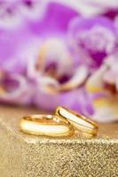Wedding gold rings on gold pedestal behind purple orchids. Vertical photo