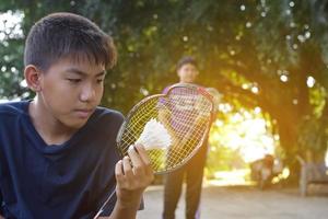 un joven asiático sostiene una raqueta de bádminton de marco roto en la mano tristemente mientras juega bádminton con su amigo fuera de la casa, concepto de bádminton al aire libre, enfoque suave y selectivo. foto