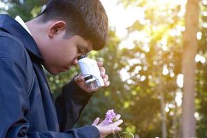 Young Asian boy is holding a pocket microscope to look at tiny particles on plant leaves to study plants and organisms on different types of plants in botanical garden, soft and selective focus. photo